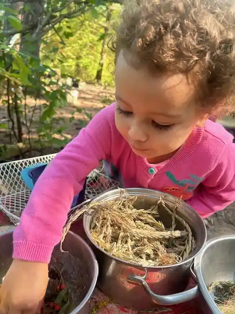 A young child with pigtails stands outdoors holding sticks near white buckets, with other children playing in the background.